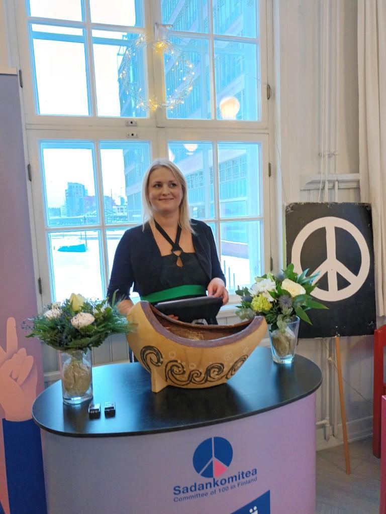 Puheenjohtaja Kaisu Kinnunen seisoo puhujanpöydän takana. Kuvassa on rauhanlaiva ja kukkia.
