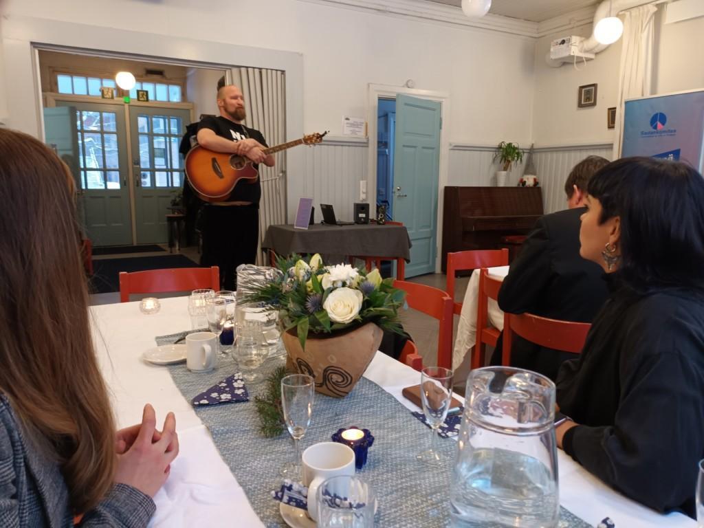Artisti Paleface soittaa kitaraa ja laulaa juhlayleisölle, jotka istuvat pöydissä. 
