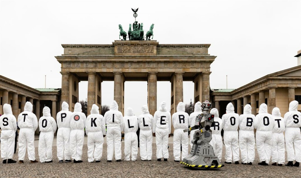 Ihmisiä seisoo rivissä selin kameraa kohti pukeutuneina valkoisiin haalareihin. Haalarien selkäpuolella olevat kirjaimet muodstavat tekstin "Stop Killer Robots".