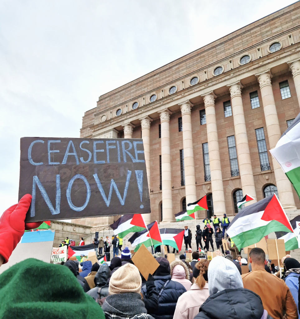 Kuvassa mielenosoittajia eduskuntatalon edessä. Etualalla kyltissä teksti "CEASEFIRE NOW!"
